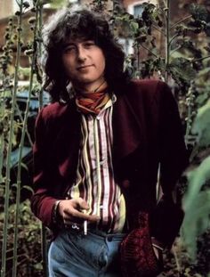 a man with long hair wearing a red jacket and striped shirt standing in front of plants