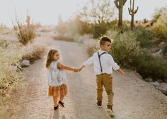 two young children holding hands and walking down a dirt road in the desert with cactus trees behind them