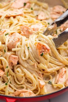 pasta with salmon and cream sauce in a red pan on a white countertop, being stirred with a pair of tongs