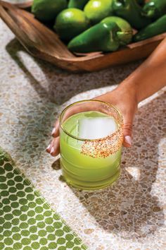 a person holding a green drink in front of a basket full of peppers and limes