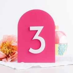 a pink table number sitting on top of a white towel next to a flower and bottle