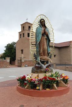 a statue of the virgin mary in front of a church with flowers on it's pedestal