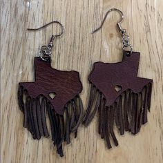 two brown leather earrings with fringes hanging from them on a wooden surface, one is shaped like a cow