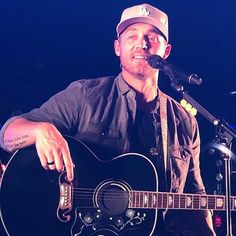 a man with a hat on holding a guitar and singing into a microphone at a concert