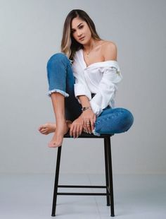 a woman sitting on top of a stool wearing blue jeans and a white off the shoulder shirt