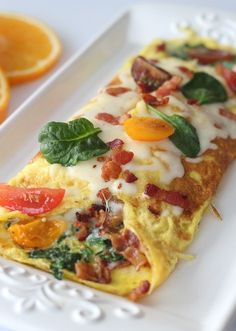 an omelet with bacon, spinach and tomatoes on a white plate next to sliced oranges