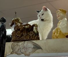 a white cat sitting on top of a shelf next to other figurines and statues