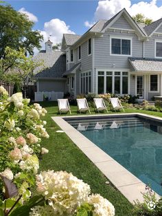 a pool in front of a large house with lawn chairs and flowers around the pool