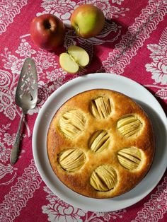 an apple pie on a white plate next to some apples and a silverware with a pink table cloth