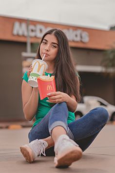 a woman sitting on the ground eating a hot dog and holding a drink in her hand