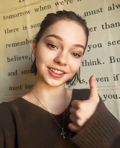 a girl giving the thumbs up sign with her hand and wearing a brown sweater, standing in front of a newspaper