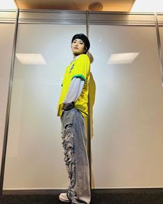 a young boy standing in front of a glass wall wearing a yellow shirt and jeans