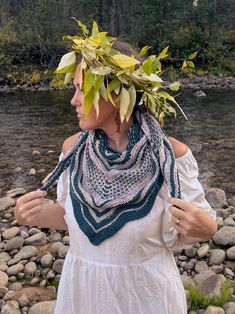 a woman in white dress standing next to water with leaves on her head and scarf around her neck