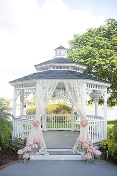 a white gazebo with pink flowers on the ground in front of it and trees