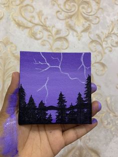 a hand holding up a purple and black painting with trees in the foreground on a white background