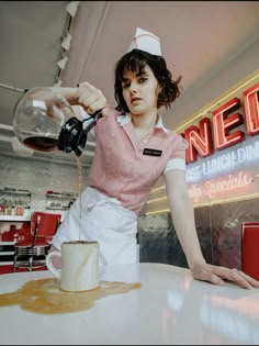 a woman pouring orange juice into a glass on top of a white table in front of a neon sign