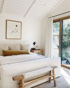 a bedroom with a large bed and white walls, along with a wooden bench in front of the window