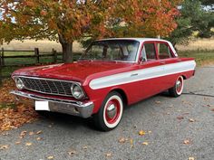 an old red and white car parked in front of a tree with leaves on the ground