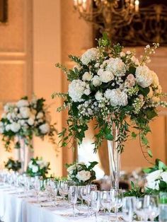 there is a table set up with white flowers and greenery in tall vases