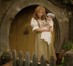 a woman holding a baby standing in front of a hobbot door with a wooden fence