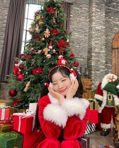 a woman sitting in front of a christmas tree wearing a santa suit and holding her hand up to her face