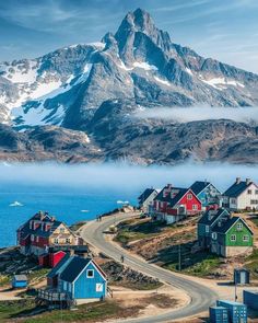 houses on the side of a mountain with snow capped mountains in the background and water below