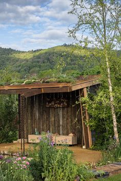 a wooden shed with a green roof surrounded by flowers and trees