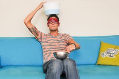 a man sitting on a blue couch holding a bowl over his head with one hand