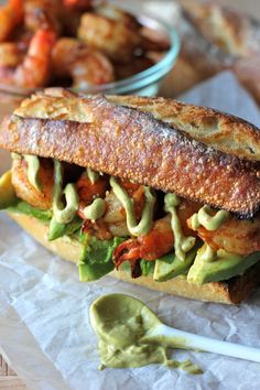 a sandwich with avocado and other toppings sits on wax paper next to a bowl of shrimp