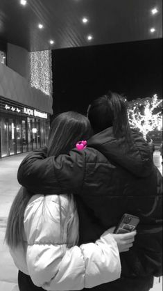 two people hugging each other in front of a building with lights on the side walk