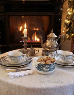 a table set for two with tea cups and plates in front of a fire place