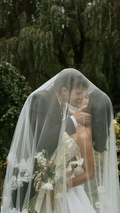 a bride and groom kissing under a veil in front of some trees on their wedding day