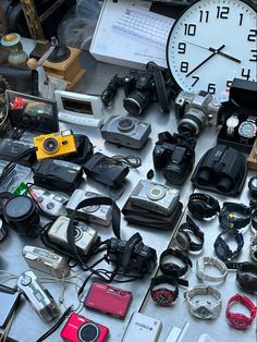 many different types of cameras and accessories on a table with a clock in the background