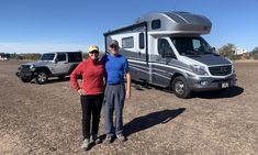 two people standing next to an rv in the middle of a dirt field with other vehicles behind them