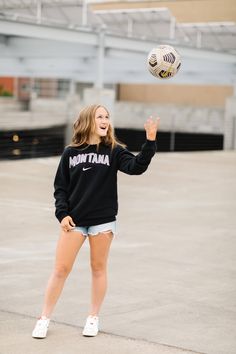 a young woman is playing with a soccer ball in an empty parking lot while wearing a black sweatshirt and white tennis shoes