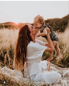 a woman holding a small child while sitting on top of a blanket in a field