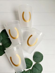 four glasses with gold foil on them sitting next to green leafy leaves and white wall