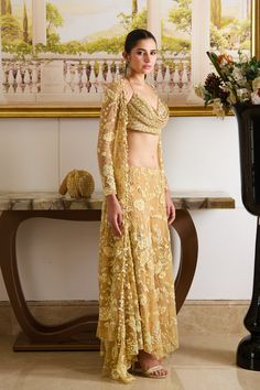 Step into the spotlight with our golden glamour co-ord set, featuring a glamorous cowl-neck top paired up with flowy pants and a complimentary full-sleeved jacket adorned with golden floral embroidery crafted out of precious sequins, crystals, and dainty detailing. The tie-up cowl neck designer blouse features intricate fish scale pattern embroidery with sequins and beads, and the heavily embellished flower motifs adorn the jacket and wide-legged flowy pants. Cowl neck blouse adorned with fish s Glamorous Floor-length Palazzo Set With Zari Work, Glamorous Designer Palazzo Set With Sheer Dupatta, Gold Pre-draped Saree With Resham Embroidery For Evening, Glamorous Palazzo Set With Traditional Drape For Parties, Glamorous Traditional Drape Palazzo Set For Parties, Elegant Floor-length Sets With Gold Embroidery, Glamorous Evening Saree Sets, Elegant Gold Embroidered Floor-length Sets, Gold Embellished Sets For Reception