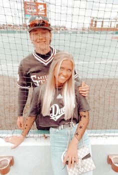 two people standing next to each other in front of a baseball field with the net behind them