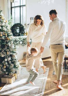 a woman and man holding hands as a toddler stands on a rug in front of a christmas tree