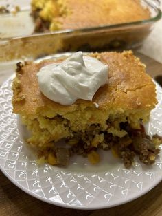 a piece of cornbread pie with sour cream on top sits on a plate next to a casserole dish