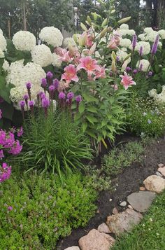 a garden filled with lots of white and pink flowers next to green grass, rocks and trees