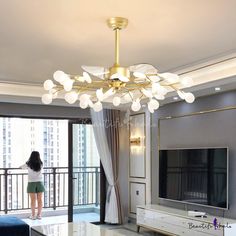 a woman standing in a living room with a tv on the wall and a large glass chandelier