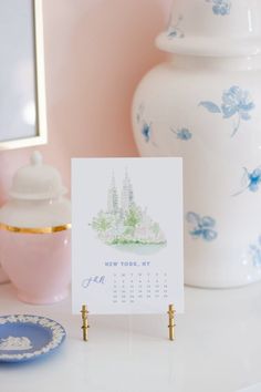 a calendar sitting on top of a table next to a white vase and pink wall