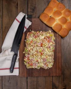 a wooden cutting board topped with bread and coleslaw next to a loaf of rolls