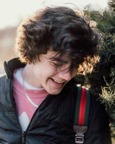 a young man with curly hair wearing a black jacket and suspenders smiling at the camera