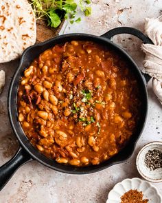 a skillet filled with beans and meat on top of a white table next to bread