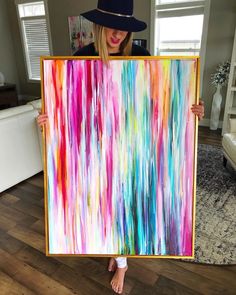 a woman holding up a colorful painting in her living room while wearing a blue hat