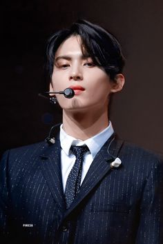 a man wearing a suit and tie with a microphone attached to his head, in front of a dark background