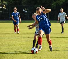 two girls playing soccer on a field with trees in the background and one girl trying to kick the ball